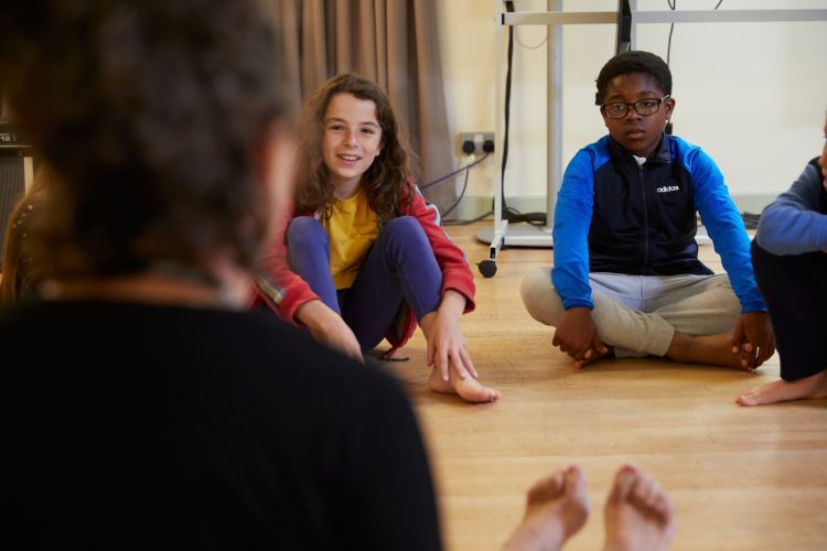 From left to right: A girl wearing a yellow top and purple leggings is sat on the floor smiling. A boy wearing a blue and black top sits next to her cross legged.